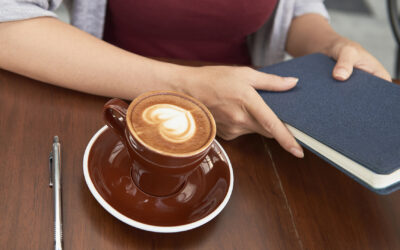 Close-up image of woman drinking cup of cappuccino and opening gratitude journal to write down her thoughts